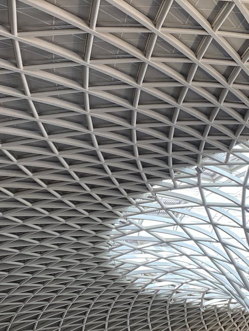 Interior view of a modern train station with a high arched ceiling made of blue steel and glass, supported by curved steel beams. The station features brick walls with arched windows, a glass railing
