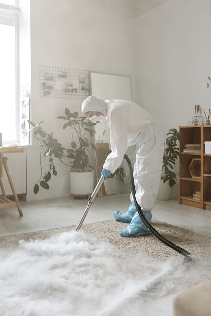 A professional cleaning technician in a white protective suit, mask, and gloves is performing surface cleaning on a beige shaggy carpet using a vacuum cleaner with a long hose attachment in a well-lit
