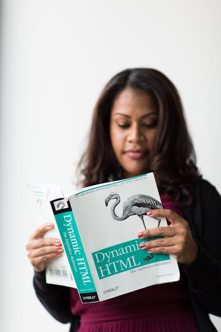 A woman with long dark hair wearing a dark blazer and magenta top is reading a book titled 'Dynamic HTML'. She is holding the book with both hands, with her right index finger pointing at the page. Th